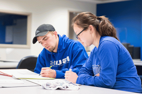 Two students, an woman with dark hair in a ponytail and glasses, and a man wearing a grey baseball cap, work on an assignment together. They both wear blue ISU apparel and both hold writing implements in their hands.