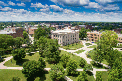 Elevated drone image of the Indiana State University campus, with Normal Hall, a white stone building, pictured in the center and other brick buildings visible nearby. The campus is shown in summer with green trees and grass visible in the foreground and around and behind the various buildings. 